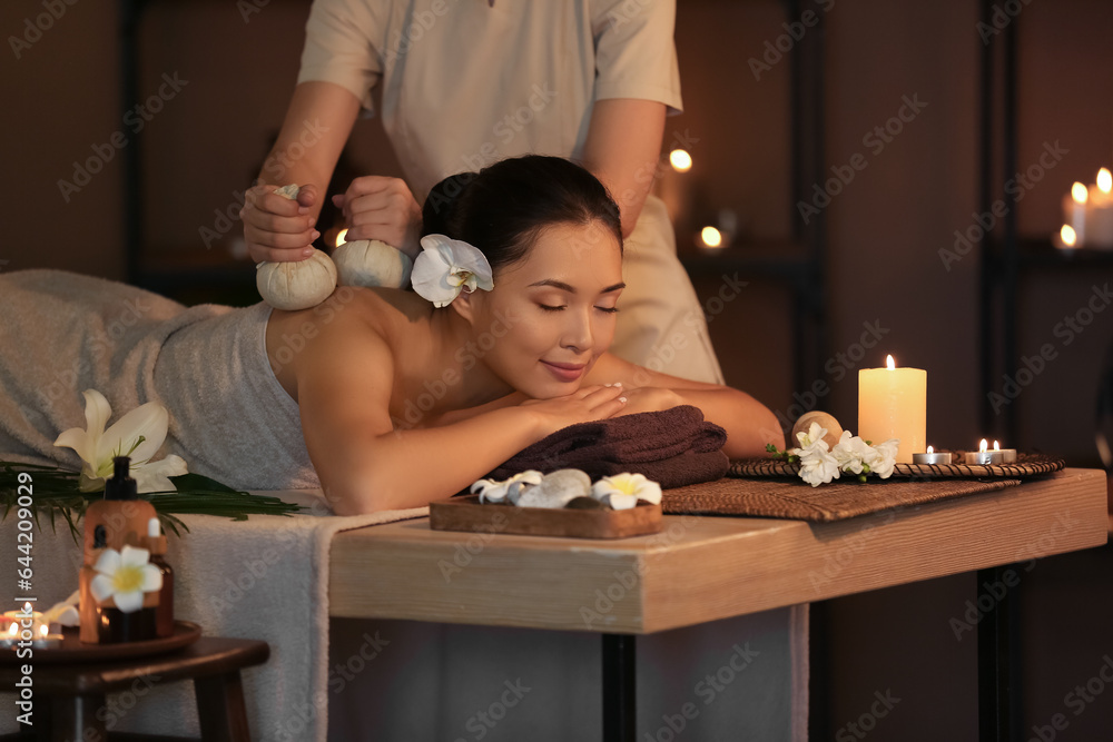 Young woman undergoing treatment with herbal bags in dark spa salon