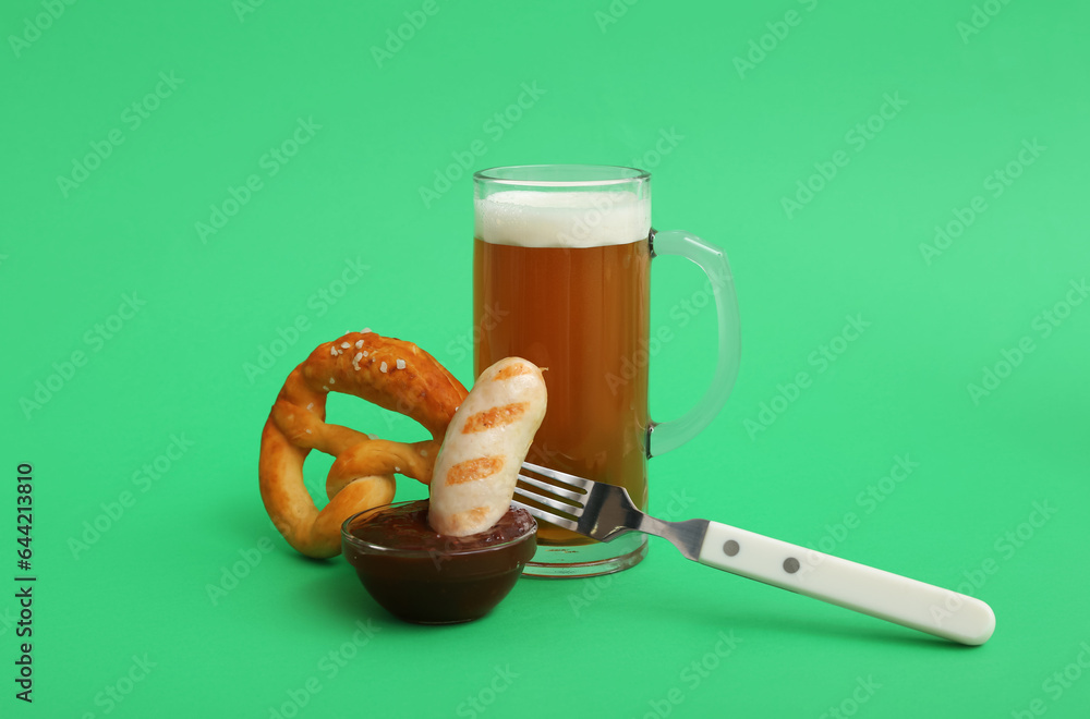 Fork with tasty Bavarian sausage, pretzel, sauce and mug of beer on green background. Oktoberfest ce