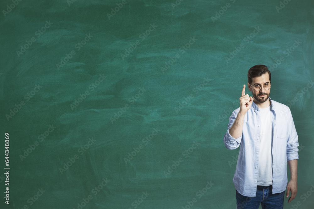 Attractive young european businessman pointing finger up while standing on empty chalkboard backgrou