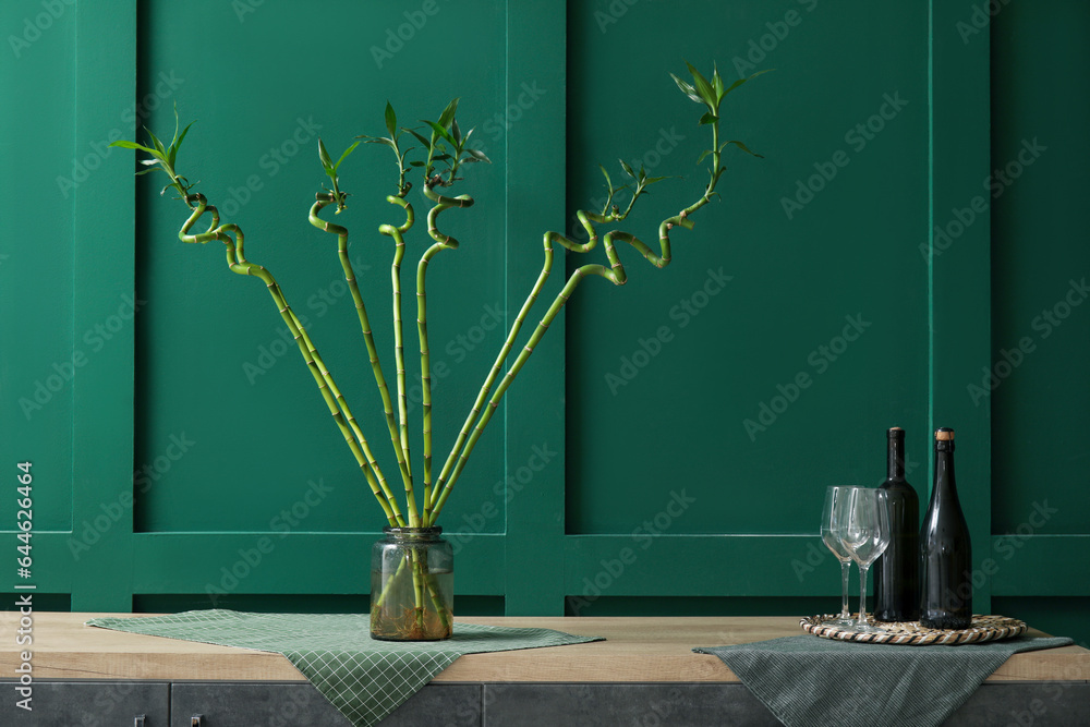 Vase with bamboo stems, bottles of wine and glasses on table near color wall