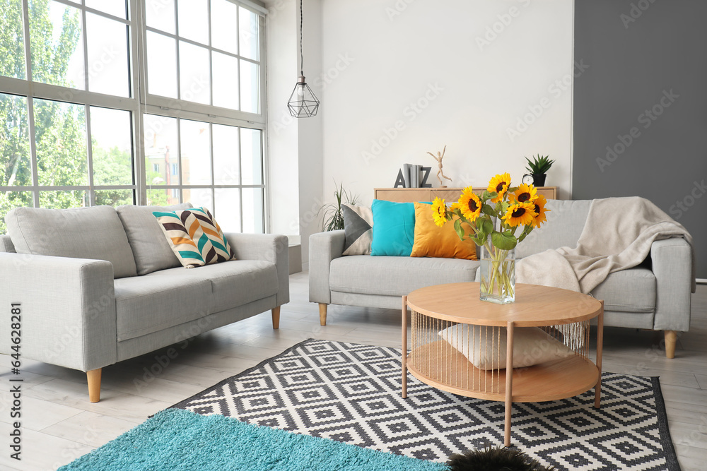 Cozy grey sofas and vase with beautiful sunflowers in interior of light living room