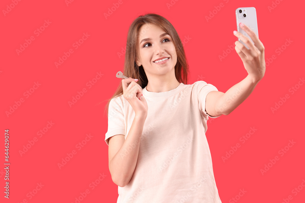 Young woman with key from house taking selfie on red background