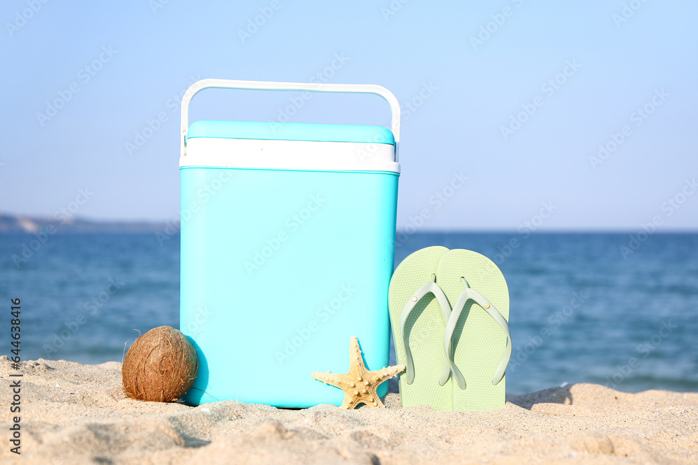 Modern beach cooler, flip-flops and ripe coconut on sand