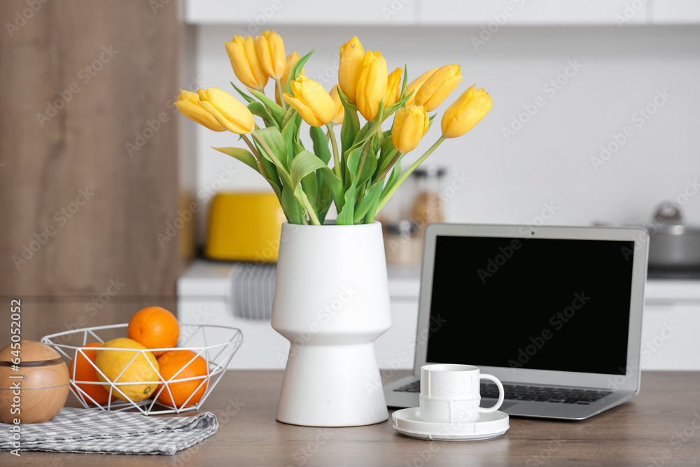 Vase with yellow tulip flowers, modern laptop, fruits and cup of coffee on wooden table in light kit