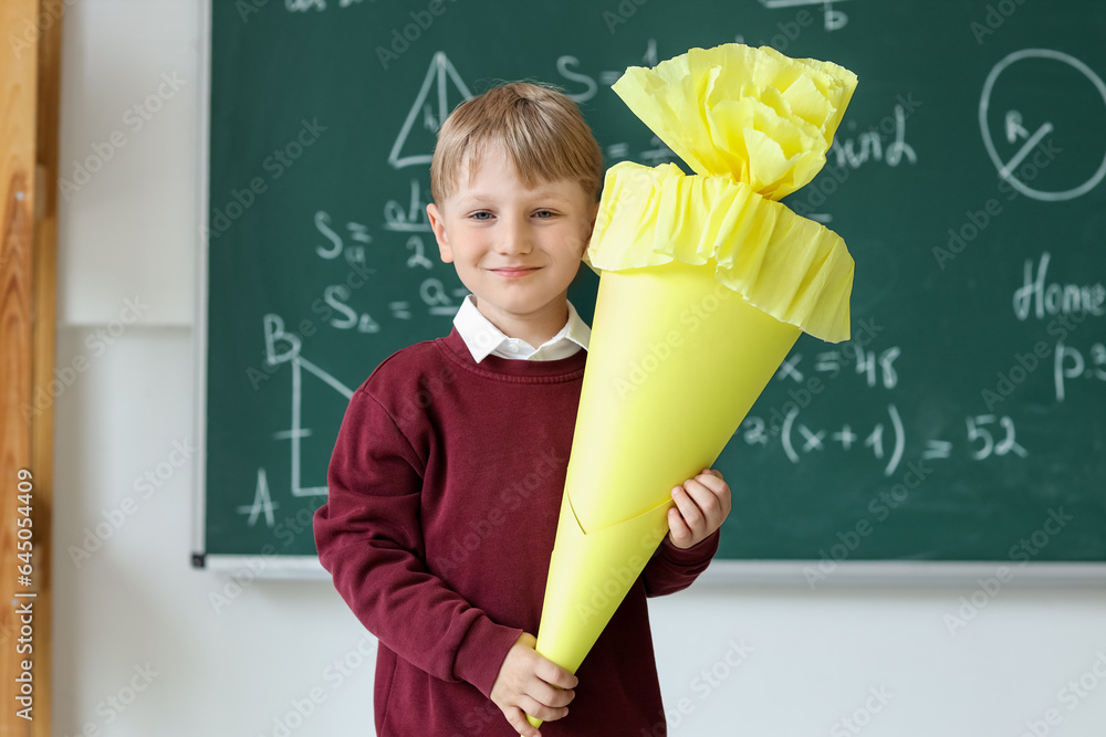 Happy little boy with yellow school cone in classroom near blackboard