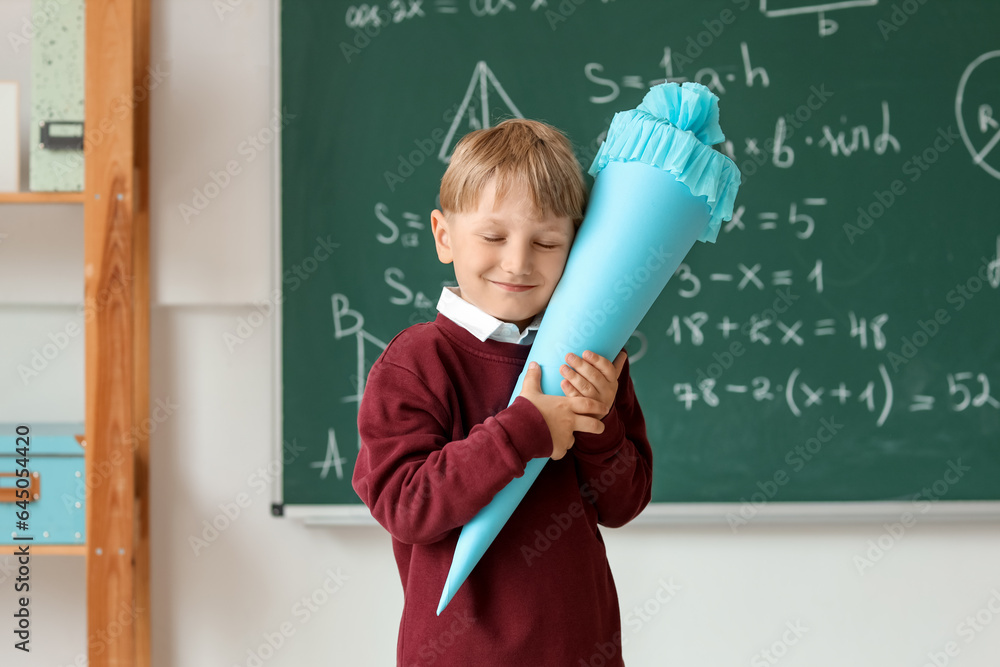 Happy little boy with blue school cone in classroom near blackboard