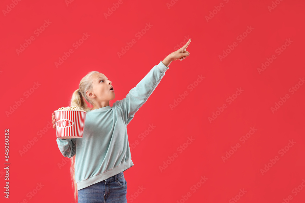 Shocked girl with bucket of popcorn pointing at something on red background