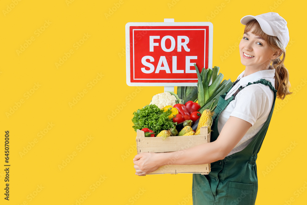 Young female farmer with wooden box full of different ripe vegetables and FOR SALE sign on yellow ba