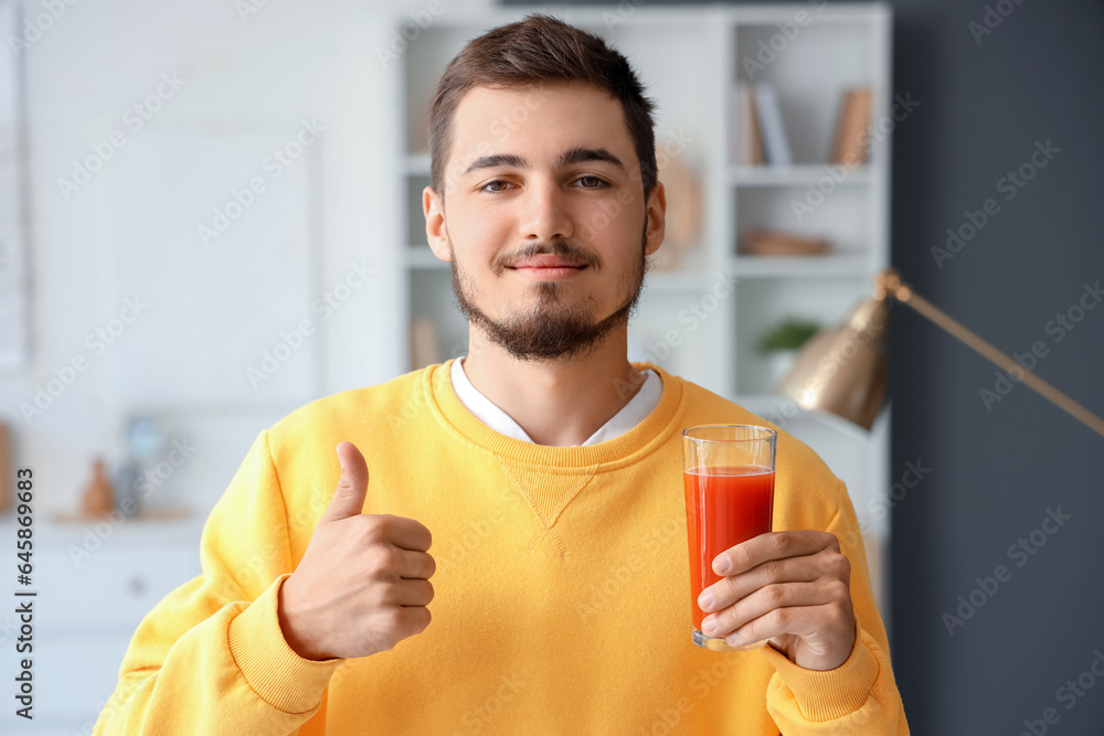 Young man with glass of tomato juice showing thumb-up at home, closeup