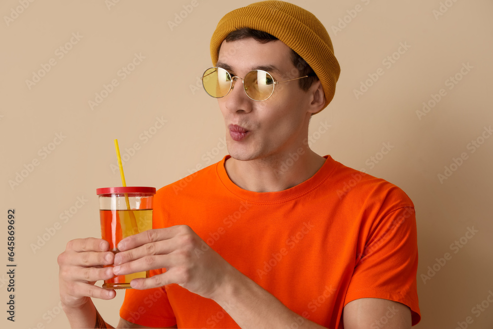 Young man with glass of juice blowing kiss on beige background, closeup