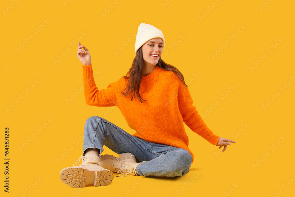 Young woman in winter clothes sitting on yellow background