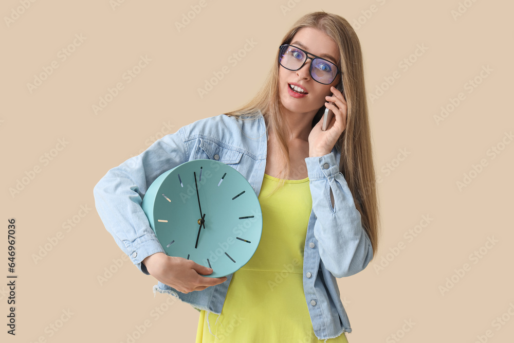 Stressed young woman with clock talking by mobile phone on beige background. Deadline concept