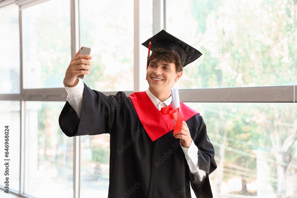 Male graduate student with diploma taking selfie near window in room