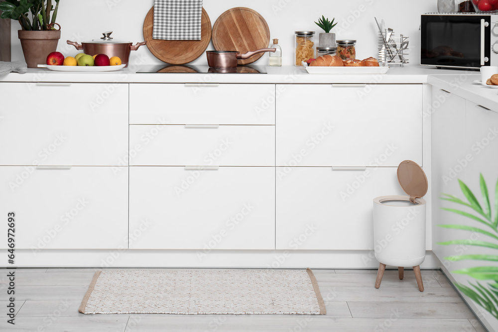 Interior of modern kitchen with stylish rug, trash bin and white counters