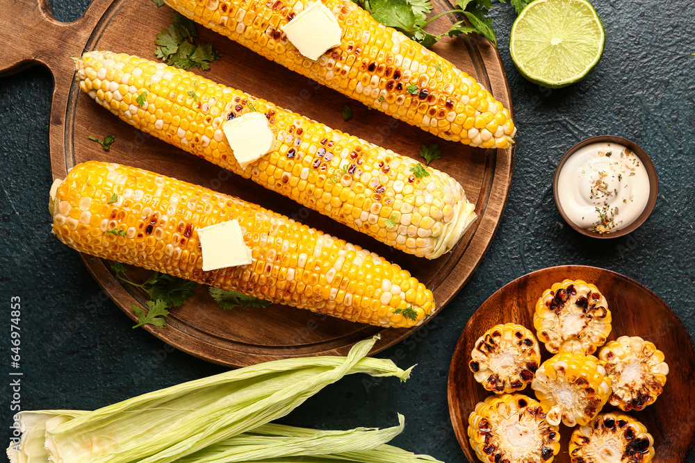 Wooden board and plate of tasty grilled corn cobs with butter on dark background