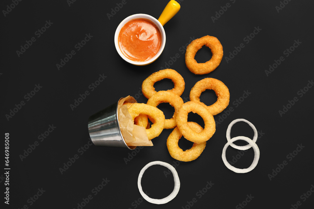 Metal bucket with fried breaded onion rings and sauce on black background