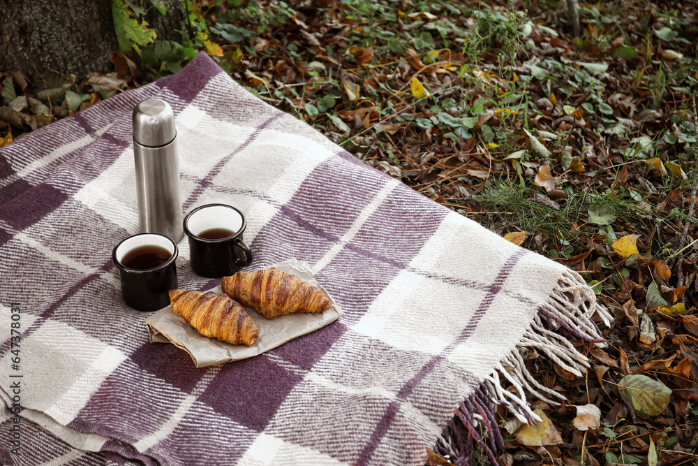 Thermos with cups of coffee and croissants on plaid in autumn park