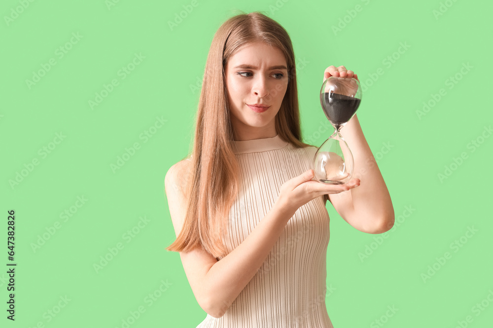 Thoughtful young woman with hourglass on green background. Deadline concept