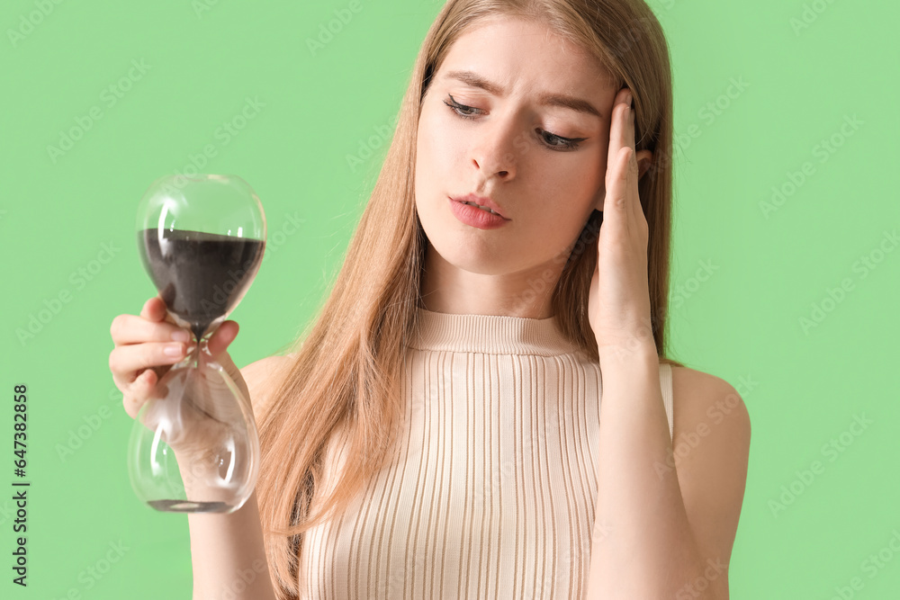 Stressed young woman with hourglass on green background, closeup. Deadline concept