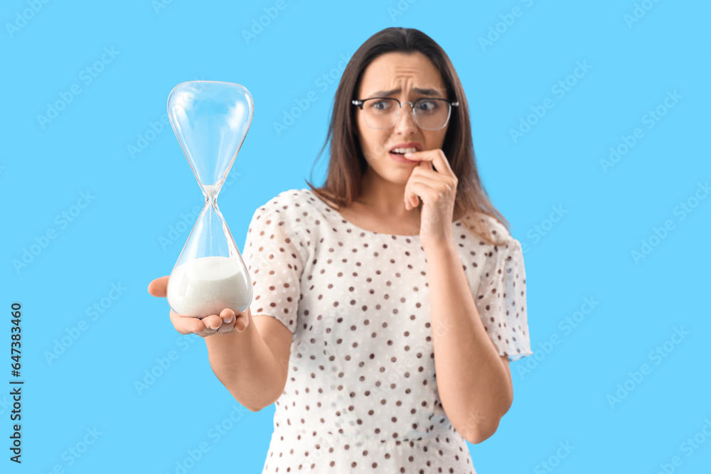 Worried young woman with hourglass on blue background, closeup. Deadline concept