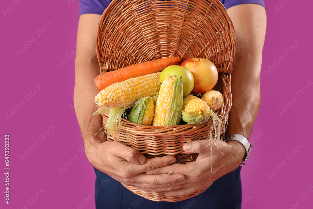 Mature male farmer with wicker basket full of ripe vegetables on purple background