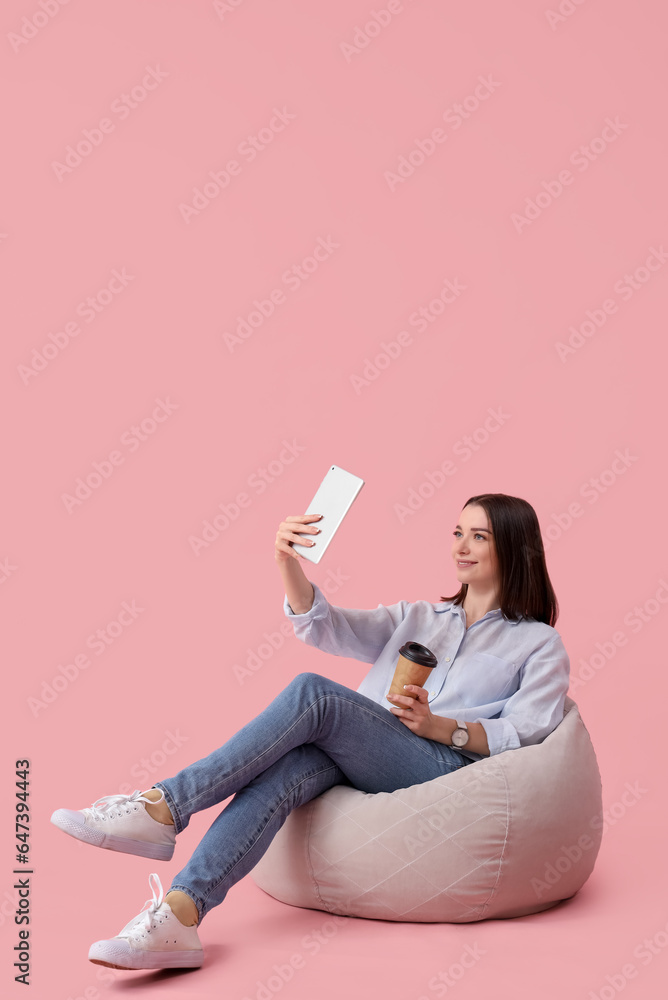 Young businesswoman with tablet computer and cup of coffee sitting on pink background