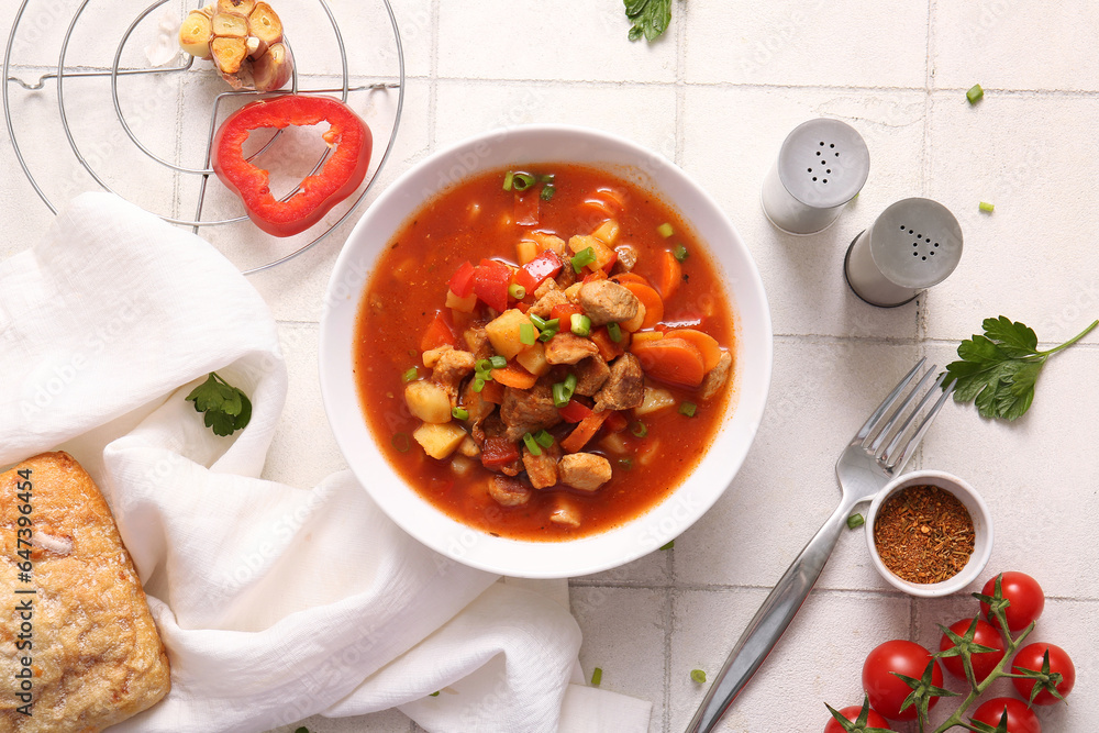 Bowl of tasty beef stew, spices and vegetables on white tile background