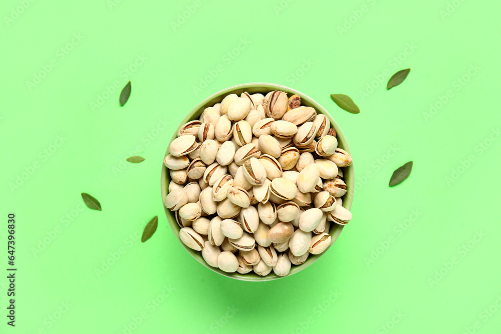 Bowl with tasty pistachio nuts and leaves on green background