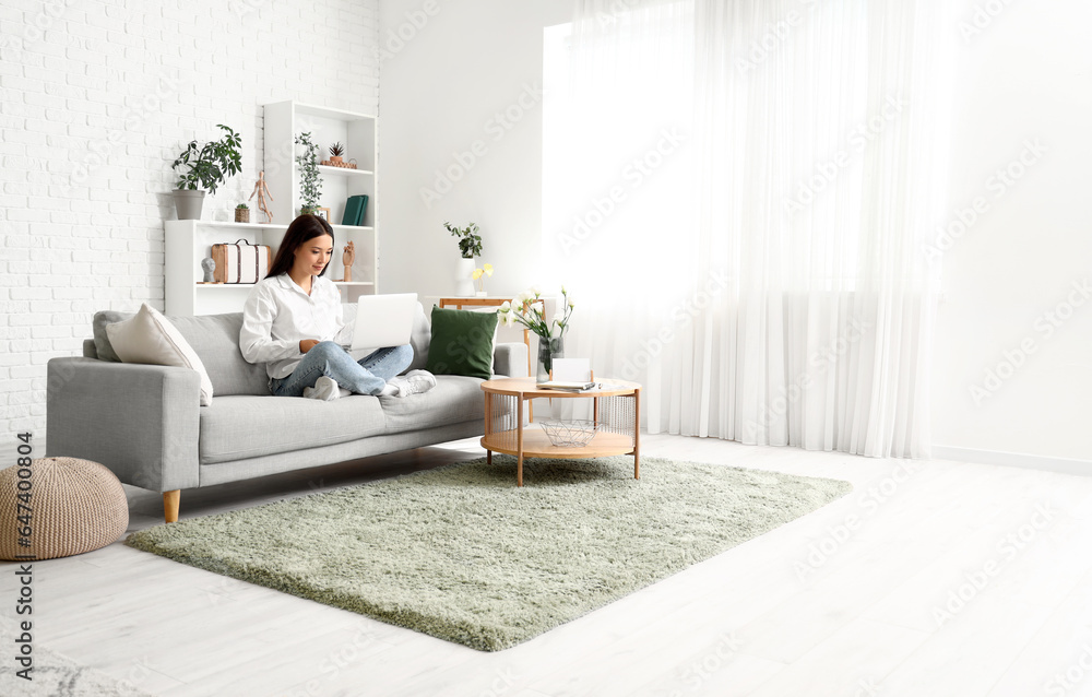 Beautiful young Asian woman with laptop sitting on sofa in living room