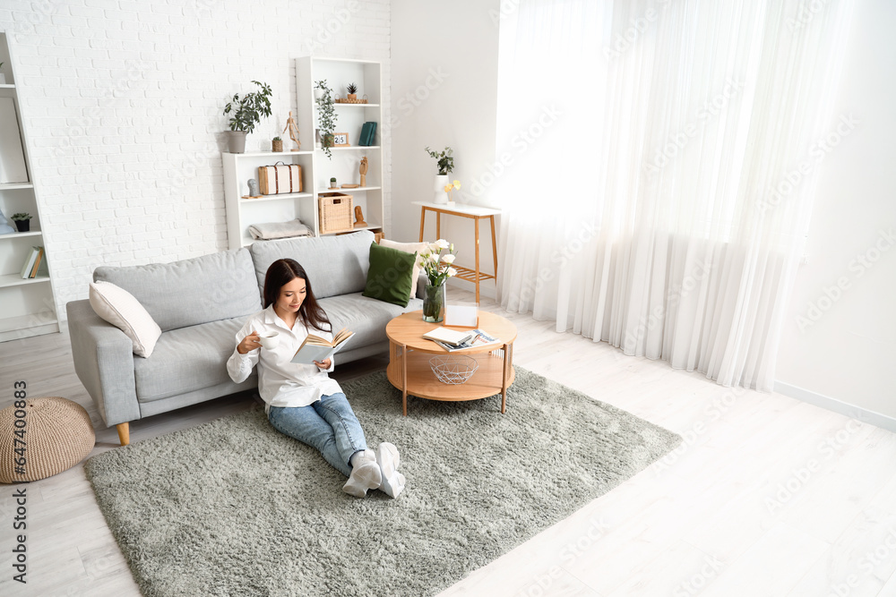 Beautiful young Asian woman with cup of tea reading book in living room