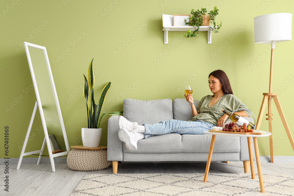 Beautiful young Asian woman with glass of wine, grapes and cheese lying on sofa in living room