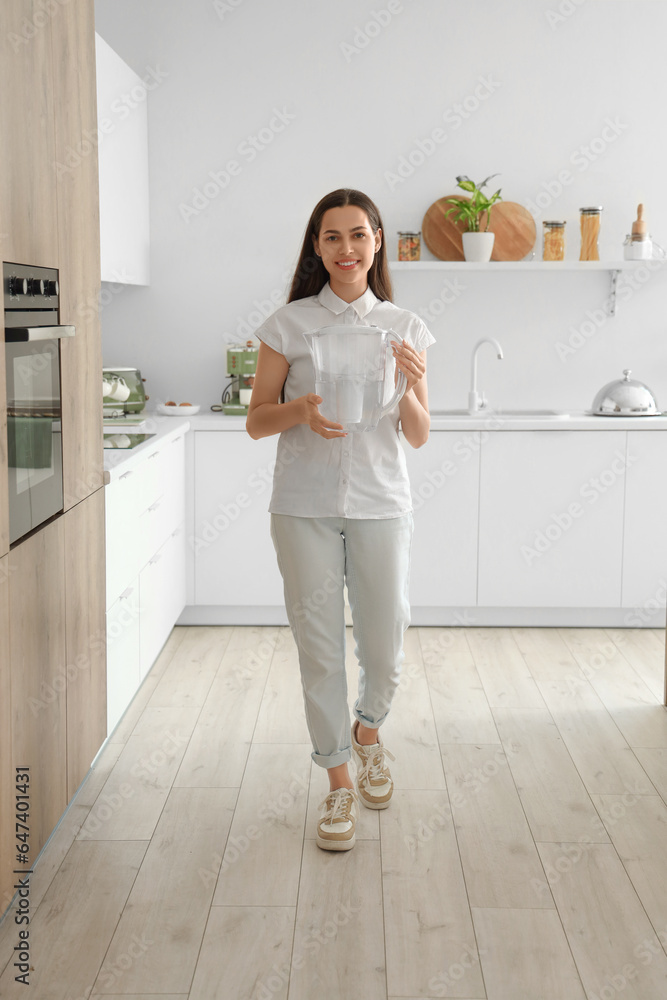 Beautiful young woman with water filter jug in kitchen