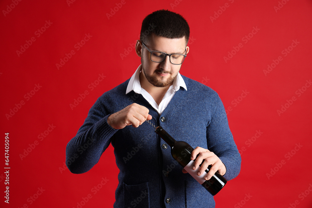 Young sommelier opening bottle of wine with corkscrew on red background