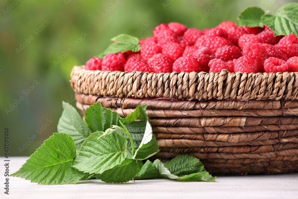 Wicker basket with tasty ripe raspberries and leaves on white wooden table against blurred green bac