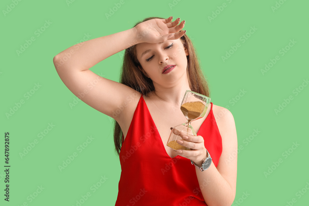 Tired young woman with hourglass on green background. Deadline concept