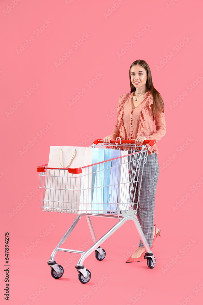 Young woman with shopping cart and bags on pink background