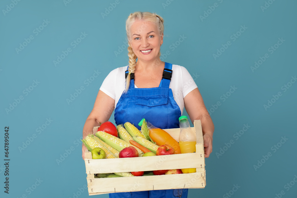 Happy mature female farmer with wooden box full of different ripe vegetables on blue background