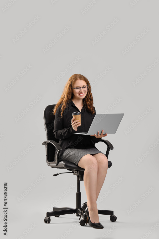 Young businesswoman with laptop and cup of coffee sitting in chair on light background