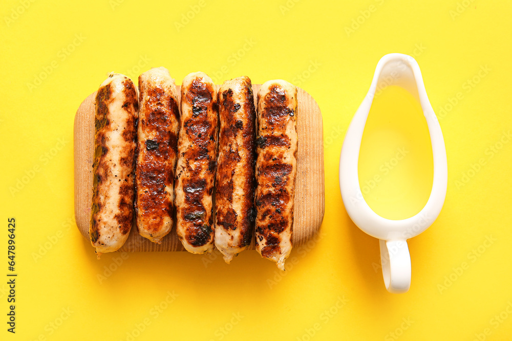 Wooden board of tasty grilled sausages and oil on yellow background