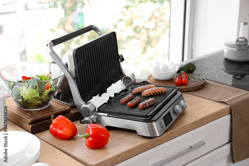 Modern electric grill with tasty sausages on wooden countertop in kitchen