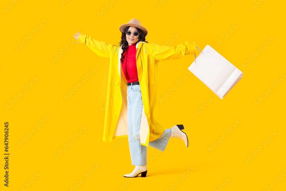 Stylish young Asian woman in fall clothes with shopping bags on yellow background