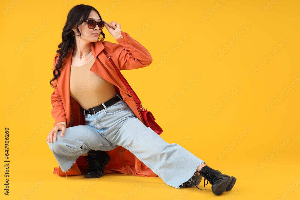 Stylish young Asian woman in fall clothes sitting on yellow background