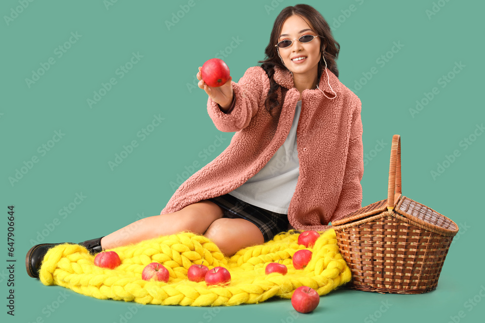 Young Asian woman in fall clothes with apples and picnic basket on green background