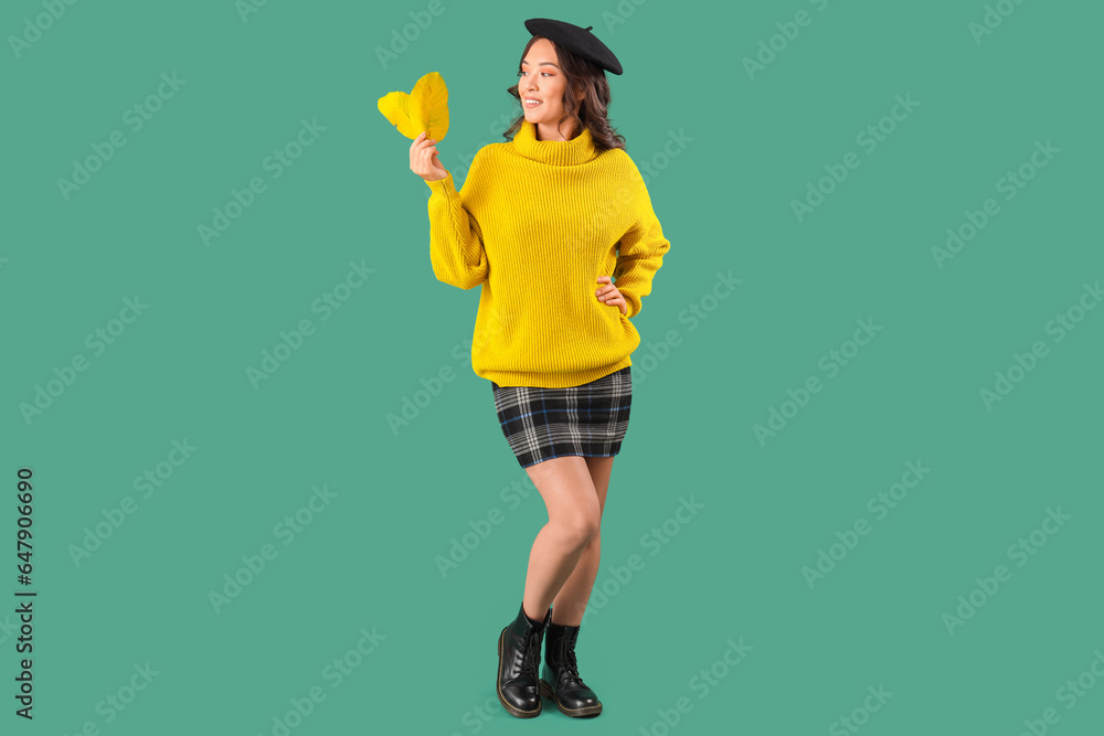 Stylish young Asian woman in fall clothes with leaves on green background