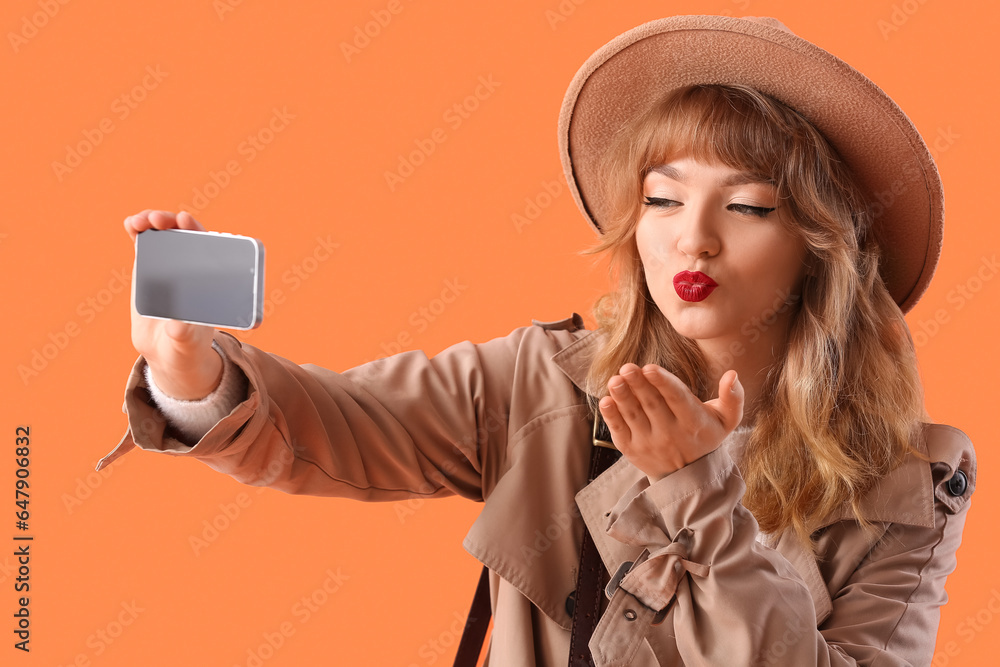 Stylish young woman in fall clothes taking selfie on orange background