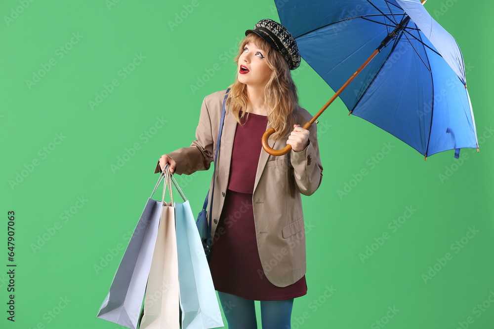 Stylish young woman in fall clothes with umbrella and shopping bags on green background