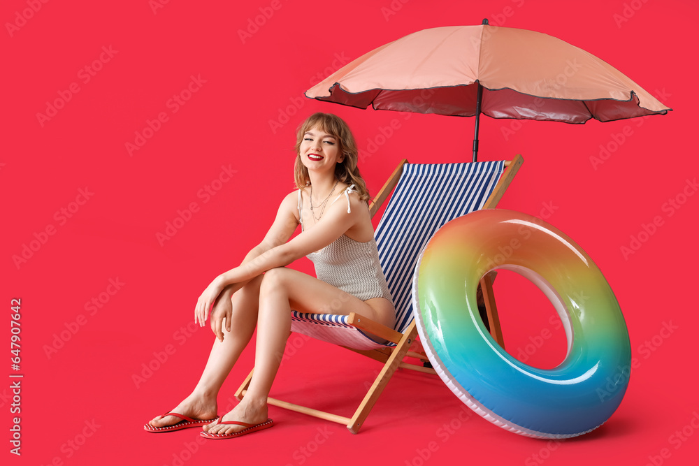 Young woman in deck chair with beach umbrella and swim ring on red background