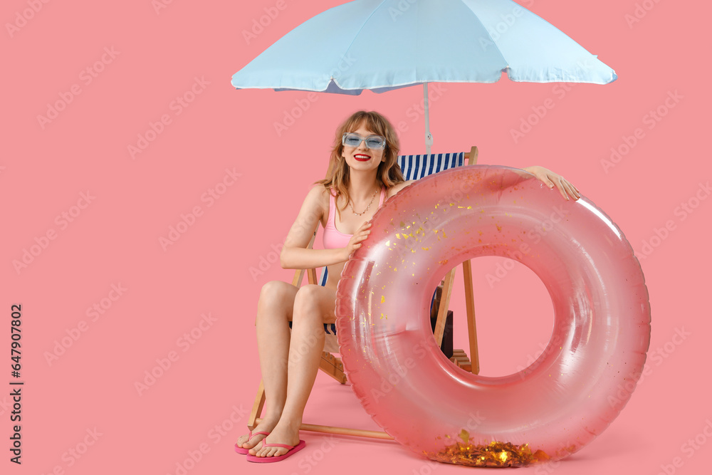 Young woman in deck chair with beach umbrella and swim ring on pink background