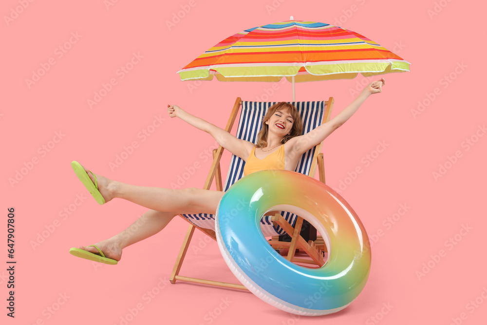 Young woman in deck chair with beach umbrella and swim ring on pink background