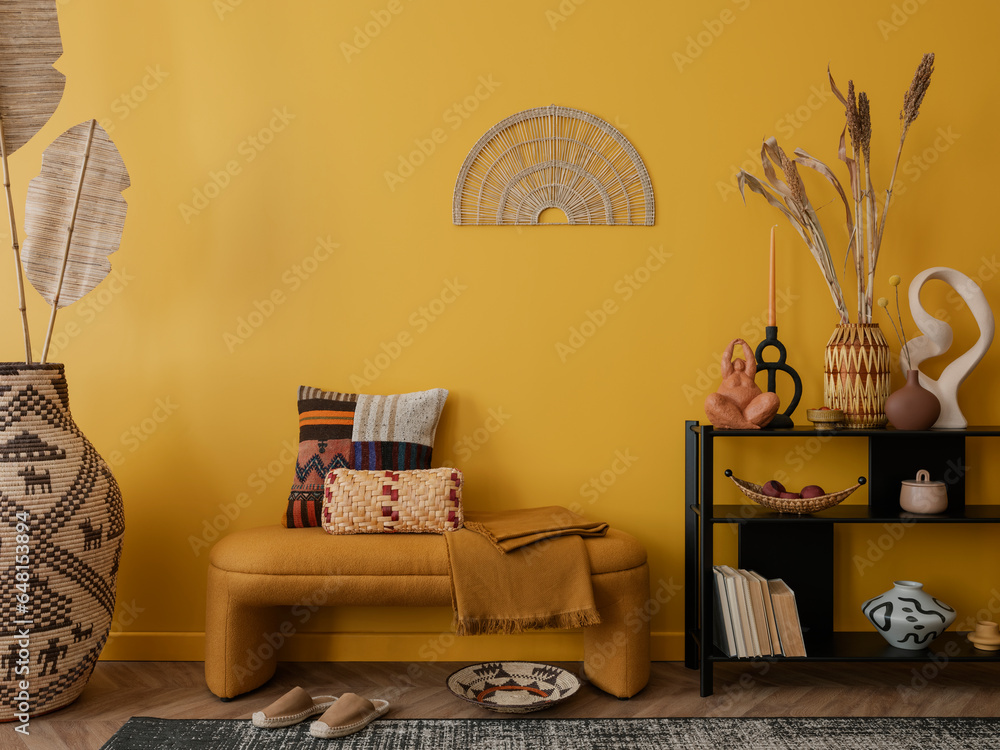 Interior design of warm living room interior with stylish bench, patterned pillows, yellow wall, ornament on wall, black rack, vase with dried flowers and personal accessories. Home decor. Template.